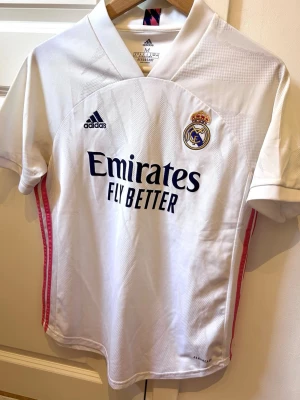 Real Madrid matchtröja Adidas vit - Snygg vit Real Madrid matchtröja från Adidas med klubbmärke och Adidas-logga på bröstet. Tröjan har svarta detaljer, rosa ränder på sidorna och 'Emirates Fly Better' tryckt framtill. Tillverkad i lätt och ventilerande material med AEROREADY-teknologi.