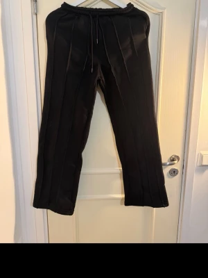 Svarta mjukisbyxor med raka ben - Säljer ett par svarta mjukisbyxor med raka ben och resår i midjan samt dragsko. Byxorna har sydda pressveck framtill för en snyggare look. Perfekta för en avslappnad och trendig stil.
