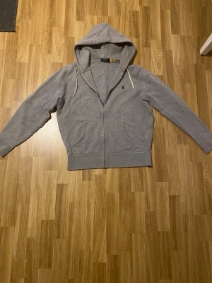 Grå hoodie från Polo Ralph Lauren - Snygg grå hoodie med dragkedja från Polo Ralph Lauren. Klassisk design med huva och vita dragsnören, två fickor framtill och den ikoniska lilla loggan broderad på bröstet. Perfekt för en avslappnad och stilren look. Kan gå ner i pris vid en snabb affär!