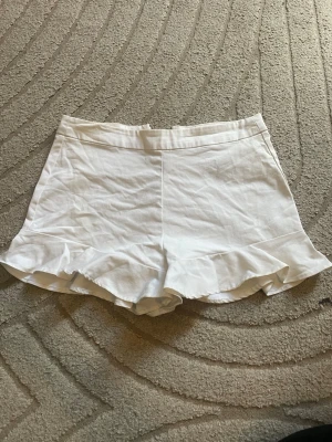 Zara Vita shorts med volangkant - Säljer ett par vita shorts med snygg volangkant nedtill. De har en enkel och clean design med hög midja och är tillverkade i ett lätt bomullsliknande material. Perfekta för varma sommardagar och ger en fräsch look.