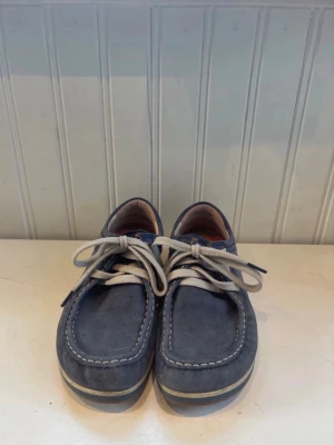 Blå mocka seglarskor med snörning - Säljer ett par blå seglarskor i mocka med vita kontrastsömmar och beige snören. Skorna har en klassisk rundad tå och låg profil, samt en bekväm innersula i orange. Perfekta för en avslappnad och stilren look. Populära!