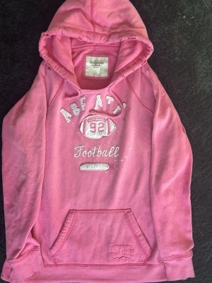 Rosa hoodie Abercrombie & Fitch - Snygg rosa hoodie från Abercrombie & Fitch med tryck framtill som visar 'A&F ATD 92 Football'. Hoodien har huva med dragsko och en stor magficka. Tillverkad i mjukt material som känns skönt mot huden. Perfekt för en avslappnad och sportig stil.