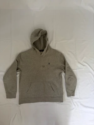 Grå hoodie från Polo Ralph Lauren - Klassisk grå hoodie från Polo Ralph Lauren med broderad svart logga på bröstet. Hoodien har en stor känguruficka framtill och mjukt material som känns skönt mot huden. Perfekt för en avslappnad och stilren look. Jätte bra skick förutom fläcken vid märket men går säkert bort i tvätten 