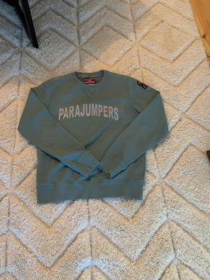 Grön sweatshirt från Parajumpers - Snygg grön sweatshirt från Parajumpers med stort tryck på bröstet och patch på ärmen. Tröjan har rund halsringning och långa ärmar. Perfekt för dig som gillar streetwear och vill ha något bekvämt och stilrent. Materialet känns mjukt och skönt mot huden. Den är typ helt ny är max använd 2 gånger men den är i barn storlek fast den största som finns så om du är runt 180 borde den passa beroende på vikt