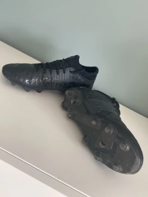 Svarta Puma Future fotbollsskor - Säljer ett par helsvarta Puma Future fotbollsskor med låg profil och stickad strumpliknande krage. Skorna har mönstrad ovandel i syntetmaterial och fasta dobbar för spel på gräs. Snygg, modern design med FUTURE-logga på sidan och klassiska snörningar.