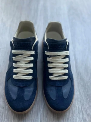 Mörkblå sneakers från Maison Margiela - Snygga mörkblå sneakers från Maison Margiela med ovandel i mocka och läder, vita snören och klassisk gummisula. Skorna har en stilren och tidlös design som passar perfekt till streetwear och casual outfits.