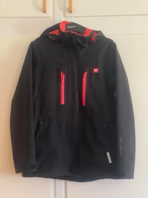 Svart Helly Hansen vindjacka med huva - Snygg svart vindjacka från Helly Hansen med röda dragkedjor och detaljer. Jackan har två bröstfickor, justerbar huva och Helly Tech-material som står emot vind och vatten. Perfekt för blåsiga dagar och outdoor-äventyr. Klassisk passform och sportig look.