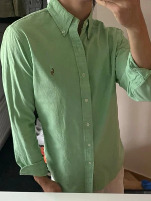 Grön skjorta från Ralph Lauren - Snygg grön skjorta från Ralph Lauren med klassisk button-down krage och broderad logga på bröstet. Skjortan har långa ärmar, normal passform och är tillverkad i mjuk bomull. Perfekt för en stilren och avslappnad look. Liten urtvättad annars bra skick 
