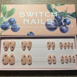 Switch Nails press-on naglar med blå blommor - Press-on naglar från Switch Nails i en ljus beige nyans med små blåbär som dekoration. Naglarna har en mandel form och kommer i flera storlekar: 0, 1, 3, 4, 8, 9, 11, 12, 13, och 14. Tillverkade av återvunnet material och levereras i en snygg förpackning med blåbärsmotiv.
