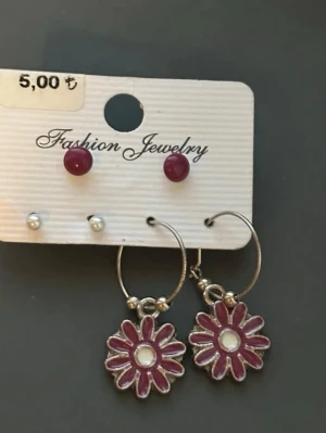 Set med örhängen med blommor och pärlor - Säljer ett set örhängen från Fashion Jewelry. Innehåller två par studs, ett par vinröda och ett par vita pärlor, samt ett par hoops med hängande blommor i vinrött och silver. Blommorna har en vit detalj i mitten och metallisk baksida. Perfekt för dig som gillar färg och söta detaljer.