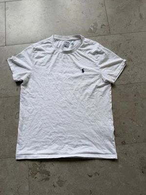 Vit t-shirt från Polo Ralph Lauren - Klassisk vit t-shirt från Polo Ralph Lauren med rund hals och korta ärmar. T-shirten har den ikoniska mörkblå broderade loggan på bröstet och är tillverkad i mjuk bomull för en skön känsla. Perfekt basic-plagg till garderoben.