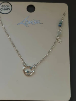 Silvrigt halsband med planet och stjärnor - Snyggt silvrigt halsband från Lovisa med en planet och små stjärnor som hänge. Kedjan är 45 cm lång och har detaljer med blå och vita pärlor samt en liten stjärnberlock på sidan. Perfekt för dig som gillar rymdinspirerade accessoarer.