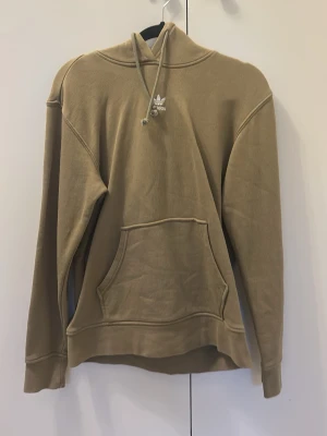 Grön hoodie från Adidas Originals - Säljer en grön hoodie från Adidas Originals med klassisk logga på bröstet. Hoodien har huva med dragsko och en stor magficka. Tillverkad i mjukt bomullsmaterial, perfekt för en avslappnad och stilren look.