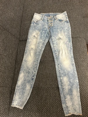 Ljusblå slitna skinny jeans - Säljer ett par ljusblå skinny jeans med slitningar och distressed detaljer på låren. Jeansen har hög midja, knappgylf och klassisk femficksmodell. Perfekta för en avslappnad och trendig look.