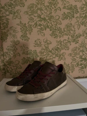 Vinröda Philippe models  - Säljer ett par bruna sneakers i skinn med vinröda om snören och detaljer i silver på hälen. Skorna har vit sula och en klassisk låg profil. Insidan är fodrad i vinrött och sidan pryds av en diskret logga. Perfekta för dig som gillar stilrena men ändå unika sneakers. Storlek 38  sulan är lite lös runt om skon