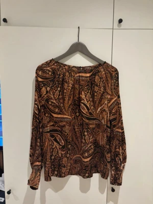 Mönstrad brun paisleyblus - Snygg blus med paisleymönster i bruna, svarta och beige toner. Blusen har långa ärmar med knappar vid ärmsluten och en avslappnad passform. Materialet känns lätt och följsamt, perfekt för att lyfta vilken outfit som helst.