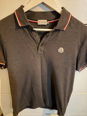 Mörkgrå Moncler pikétröja  - Snygg mörkgrå pikétröja från Moncler med klassisk krage och knappar framtill. Tröjan har röda och vita detaljer på krage och ärmslut samt Moncler-logga på bröstet. Perfekt till jeans eller shorts för en clean look.