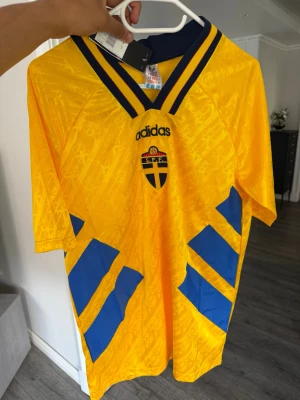 Sverige matchtröja Adidas gul/blå - Klassisk gul matchtröja från Adidas med blå detaljer och Sveriges landslagsemblem på bröstet. Tröjan har V-ringad krage med blå och gula ränder samt stora blå fält på sidorna. Tillverkad i lätt och ventilerande polyester, perfekt för fotboll eller träning.
