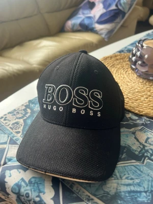 Snygg Hugo boss keps - Snygg svart keps från Hugo Boss med stor vit BOSS-logga broderad framtill. Kepsen har böjd skärm, justerbart spänne bak och är tillverkad i ett ventilerande mesh-liknande material. Perfekt för en stilren och sportig look.