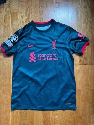 Liverpool Nike bortatröja blå - Snygg Liverpool FC bortatröja från Nike i mörkblått med röda detaljer. Tröjan har tryckta loggor, Champions League-märke på ärmen och sponsortryck framtill. Tillverkad i lätt och ventilerande material, perfekt för fotboll eller träning.