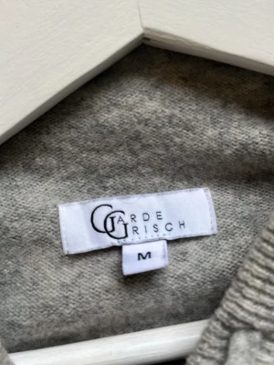 Grå stickad hoodie från Garde Grisch - Mysig grå stickad hoodie från Garde Grisch i storlek M. Tröjan har huva, långa ärmar och ribbstickade muddar vid ärmslut och nederkant. Perfekt för dig som gillar en enkel och stilren look. Materialet känns mjukt och bekvämt. Tröjan är i otroligt bra skick då den är knappt använd. Nypris:1199kr