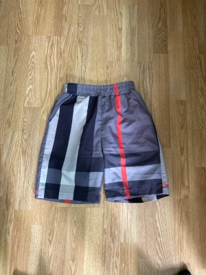 Randiga shorts med resårmidja - Snygga shorts med breda ränder i svart, grått, vitt och en röd accent. De har elastisk midja för skön passform och är tillverkade i ett lätt, syntetiskt material. Perfekta för varma dagar när du vill ha en avslappnad men trendig look.