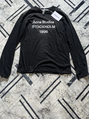 Svart långärmad topp Acne Studios - Svart långärmad topp från Acne Studios med tryck 'Acne Studios STOCKHOLM 1996' på bröstet. Tillverkad i mjukt material med rund hals och enkel, stilren design. Perfekt för dig som gillar minimalistisk och trendig stil.