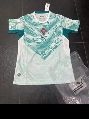 Portugal matchtröja mintgrön Puma - Snygg Portugal matchtröja från Puma i mintgrönt och vitt med grafiskt mönster. Tröjan har korta ärmar, rund hals och Portugal-emblemet på bröstet. Tillverkad i lätt och ventilerande material, perfekt för träning eller match.