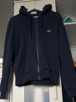 Mörkblå zip hoodie från Lacoste - Mörkblå hoodie från Lacoste med dragkedja framtill och klassisk krokodil-logga på bröstet. Tröjan har huva med dragsko, två fickor och ribbade muddar. Perfekt för en chill och stilren look. Materialet känns mjukt och bekvämt.