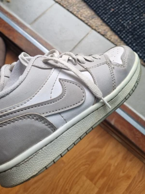 Nike Dunk Low grå/vit sneaker - Snygga Nike Dunk Low sneakers i grått och vitt med klassisk Swoosh på sidan. Skon har en låg siluett, vita paneler i textil och gråa partier i läder. Yttersulan är vit med grått mönster. Perfekt för dig som gillar stilrena och tidlösa sneakers.