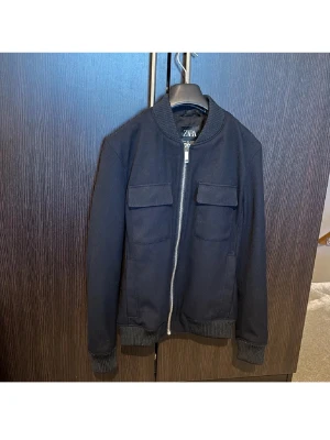 Svart bomberjacka från Zara - Snygg svart bomberjacka från Zara med ribbade muddar vid ärmslut och nederkant. Jackan har två bröstfickor med lock och dragkedja framtill. Klassisk bombermodell med rundad krage och stilren look. Perfekt för dig som gillar enkel och clean stil.