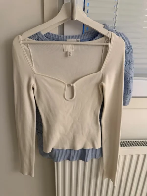 Vit långärmad topp från H&M - Trendig vit långärmad topp från H&M med snygg v-ringning och dekorativ metallring framtill. Toppen är ribbad och har en slim passform som framhäver figuren. Perfekt att styla med jeans eller kjol för en cool look.