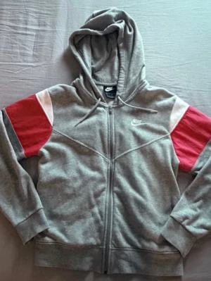 Grå Nike hoodie  - Snygg grå hoodie från Nike med dragkedja och huva. Ärmarna har färgblock i rött och vitt. 