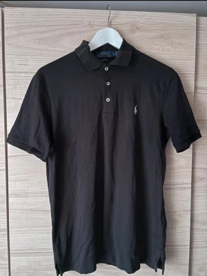 Svart polotopp från Polo Ralph Lauren - Klassisk svart pikétröja från Polo Ralph Lauren med korta ärmar och krage. Tröjan har tre knappar framtill och den ikoniska broderade loggan på bröstet. Golfpike