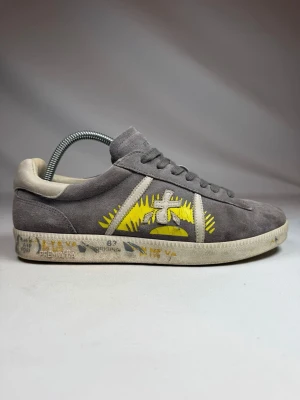 Grå Premiata sneakers i mocka - Snygga grå sneakers från Premiata i mjuk mocka med beige detaljer och coolt gult och vitt grafiskt tryck på sidan. Skorna har klassisk låg profil, grå snörning och unik printad sula. Perfekta för dig som gillar streetstyle och vill sticka ut.