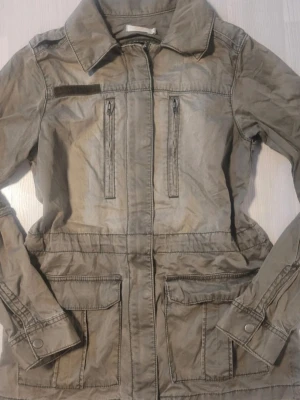 Vintage militär-stil jacka - Olivgrön jacka med dragkedja framtill. Jackan har krage och justerbara ärmslut med tryckknapp. Man kan dra på snörena på insidan för midjan! Materialet är tunt ganska och passar perfekt för våren och sommaren ifall det är lite blåsigt. Storleken är M men på grund av att den är gammal så tror jag att de passar bäst till S eller även XS 