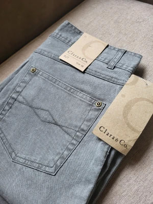 Gråa bootcut jeans Clara & Co 38 - Snygga gråa bootcut jeans från Clara & Co med klassisk femficksdesign och detaljer i metall. Jeansen har hög midja och är tillverkade i ett mjukt bomullsmaterial med lite stretch för extra komfort. Perfekta för dig som gillar en retroinspirerad look.