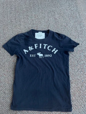 Svart t-shirt från Abercrombie & Fitch - Klassisk svart t-shirt från Abercrombie & Fitch med vit logga och text 'A&FITCH EST 1892' framtill. T-shirten har rund hals och korta ärmar, perfekt för en avslappnad och stilren look.