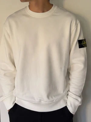 Vit sweatshirt från Stone Island - Säljer en clean vit sweatshirt från Stone Island med klassisk rund hals och ikonisk patch på vänster ärm. Tillverkad i mjuk bomull som känns skön mot huden. Perfekt för dig som gillar minimalistisk stil och streetwear-vibe.