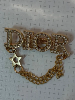 Dior brosch med kedjor och stjärna - Snygg brosch med gnistrande stenar på bokstäverna. Broschen har två hängande guldfärgade kedjor och en dekorativ stjärna. 🌟 Perfekt att sätta antingen på väskan / kläderna eller kanske till och med bikinin i sommar? 😍👌🏼