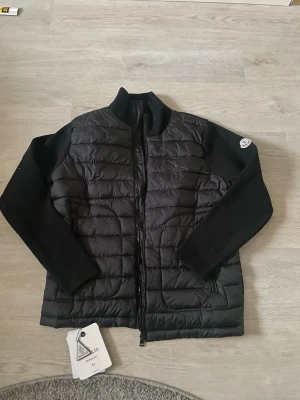 Svart cardiganjacka från Moncler - Snygg svart cardiganjacka från Moncler med quiltad front och stickade ärmar. Jackan har dragkedja framtill, hög krage och Moncler-logga på sidan 