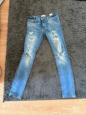 Blå slitna skinny jeans från Jack and Jones  - Säljer ett par blå skinny jeans från Jack and Jones  med flera slitningar och hål på framsidan. Jeansen har klassisk femficksdesign, normal midja och är tillverkade i stretchigt denimtyg för en bekväm passform. Perfekta för en trendig och avslappnad look.