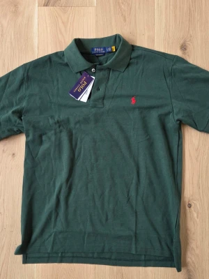 Grön pikétröja från Polo Ralph Lauren - Mörkgrön pikétröja från Polo Ralph Lauren med klassisk krage och knappar framtill. Tröjan har det ikoniska röda logobroderiet på bröstet och är tillverkad i mjuk bomull. Perfekt för en clean och stilren look.