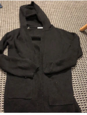 Svart hoodiekofta från KappAhl - Mysig svart kofta med huva från KappAhl. Koftan har långa ärmar, två stora fickor framtill och är tillverkad i ett mjukt, stickat material. Perfekt för lager på lager och chill dagar.