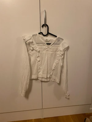 Vit blus med volanger från H&M - Söt vit blus från Gina Tricot med långa ärmar och volangdetaljer framtill. Blusen har en rundad halsringning och spetsinslag upptill. Tillverkad i ett lätt, luftigt tyg som ger en romantisk känsla. Perfekt att styla med jeans eller kjol. Inga defekter storlek 152