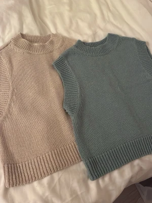 Stickade västar i beige och blågrå - Två stickade västar i mjukt garn, en i beige och en i blågrå. Båda har ribbstickad hög hals och breda muddar vid ärmslut och nederkant. 80kr/styck 