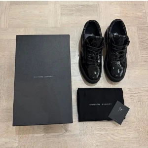 Svarta sneakers från Giuseppe Zanotti - Säljer ett par svarta sneakers från Giuseppe Zanotti i blankt lackskinn med snörning och dragkedjor på sidorna. Skorna har rund tå, platt sula och coola detaljer som logotyp på plösen och häl. Perfekt för dig som gillar statement och lyxig streetstyle.