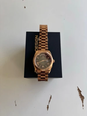 Guldfärgad klocka från Pagani Design - Stilren klocka från Pagani Design i guldfärgad metall med svart urtavla och romerska siffror. Länken är klassiskt designad och boetten har räfflad kant. Klockan har datumvisning och en elegant look som passar till många outfits. 