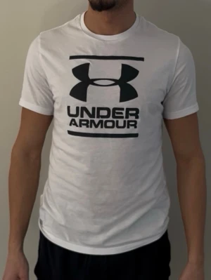  Under Armour t-shirt  - Säljer en vit t-shirt från Under Armour med stor svart logga och text på bröstet. T-shirten har korta ärmar och en klassisk passform. Perfekt för dig som gillar sportig och enkel stil.