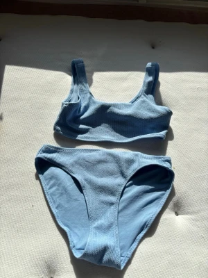 Ljusblå bikini från H&M - Säljer en ljusblå bikini från H&M med sportig topp och klassiska bikinitrosor. Bikinin har breda axelband och är tillverkad i ett ribbat syntetmaterial som ger en modern och bekväm känsla. Perfekt för lata dagar på stranden eller poolen.